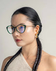 A woman with long, dark hair styled in a braided ponytail wears black cat-eye Perla II glasses by For Art's Sake® featuring blue-tinted lenses and 18-karat gold plating. She has gold hoop earrings and is dressed in a knitted beige top while standing against a plain light-colored background.