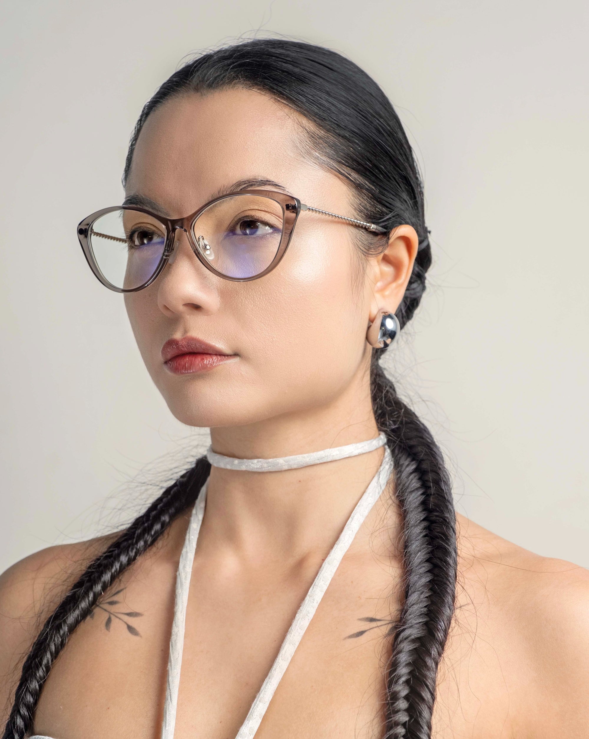 A person with long dark hair styled in two braided pigtails and wearing For Art&#39;s Sake® Perla II glasses with a thin frame featuring jade-stone nose pads. The individual has a neutral expression and wears a choker-style necklace and earrings, with small tattoos visible on their shoulders. The background is plain.