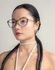 A person with long dark hair styled in two braided pigtails and wearing For Art's Sake® Perla II glasses with a thin frame featuring jade-stone nose pads. The individual has a neutral expression and wears a choker-style necklace and earrings, with small tattoos visible on their shoulders. The background is plain.