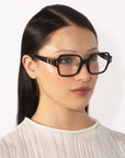 A person with long, straight dark hair is wearing large square glasses from For Art's Sake®'s Hart collection and a white pleated blouse. The plain white background highlights the individual's features and attire.