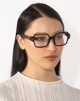 A woman with long, straight black hair is wearing oversized, handcrafted Hart eyewear by For Art's Sake®, featuring square glasses. She has a calm expression and is dressed in a light-colored, pleated blouse against a plain white background.