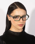 A woman with long, straight black hair is wearing large, hexagonal-shaped glasses from For Art's Sake® called Hart, featuring thick, dark green frames. She is dressed in a black, high-neck top and is facing the camera with a neutral expression against a plain white background.