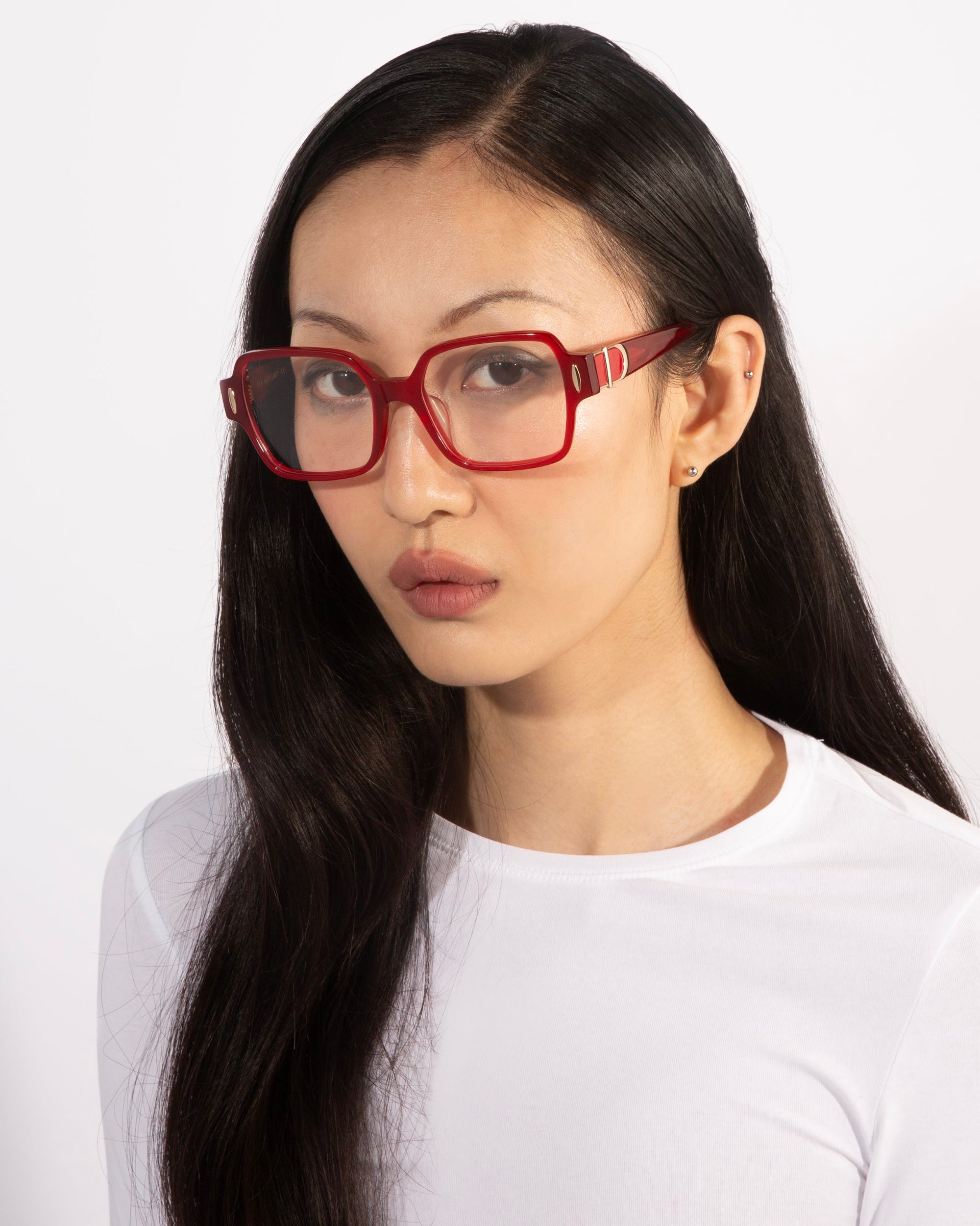 A woman with long black hair wearing the bold red square Hart glasses from For Art&#39;s Sake®, paired with a white top. She has a neutral expression and stands against a plain white background, showcasing a classic design.