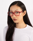 A woman with long black hair wearing the bold red square Hart glasses from For Art's Sake®, paired with a white top. She has a neutral expression and stands against a plain white background, showcasing a classic design.