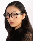 A person with long, dark hair, wearing square glasses from For Art's Sake®'s Hart collection and a black lace top, looks at the camera against a plain white background. The individual has a serious expression and their hair is styled smoothly.