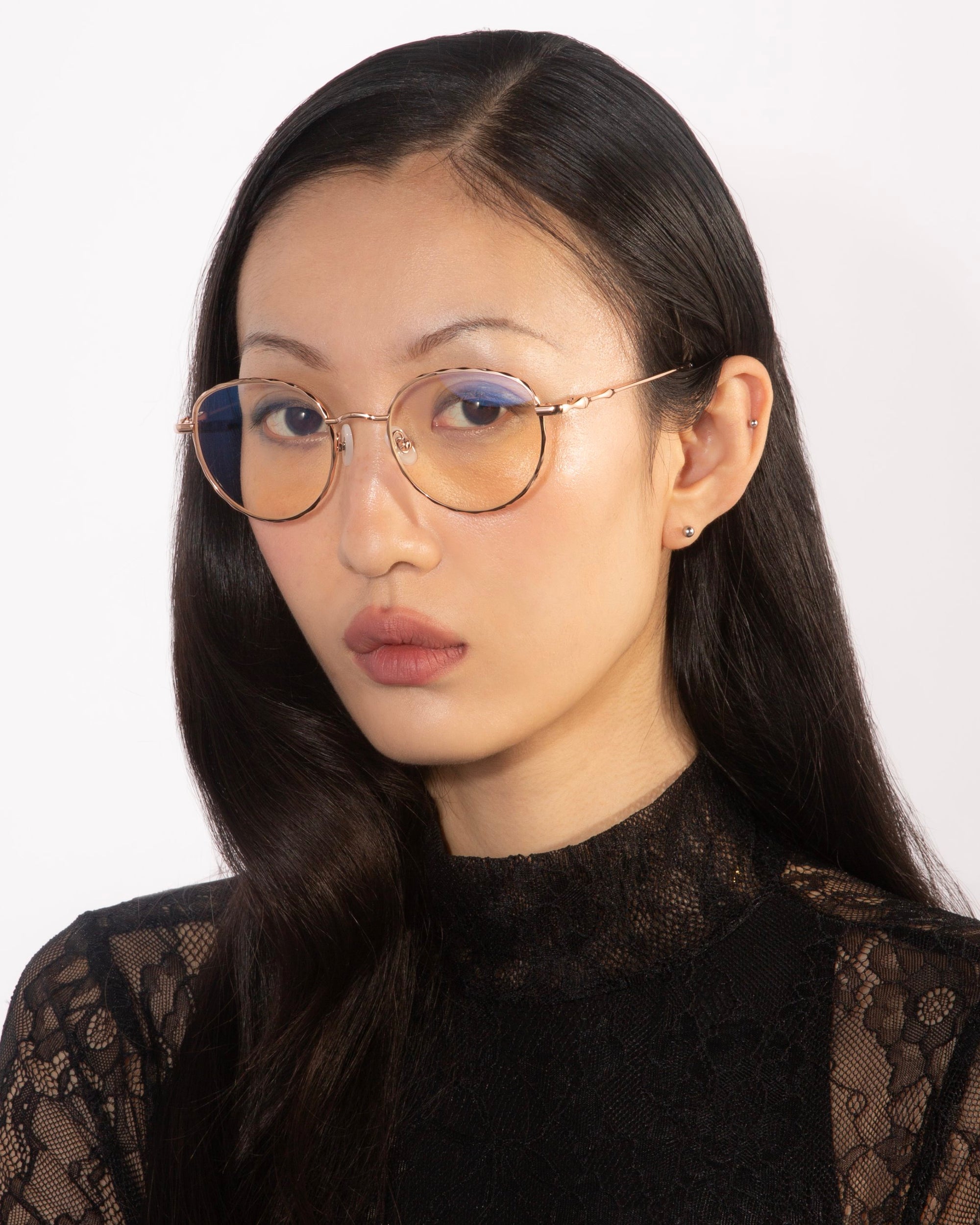 A woman with long dark hair is wearing the Bone I glasses from For Art's Sake®. The glasses have round, gold frames with a blue tint and jade stone nose pads. She has a neutral expression and is dressed in a black lace top, set against a plain white background.