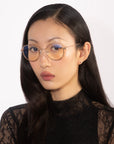 A woman with long dark hair is wearing the Bone I glasses from For Art's Sake®. The glasses have round, gold frames with a blue tint and jade stone nose pads. She has a neutral expression and is dressed in a black lace top, set against a plain white background.