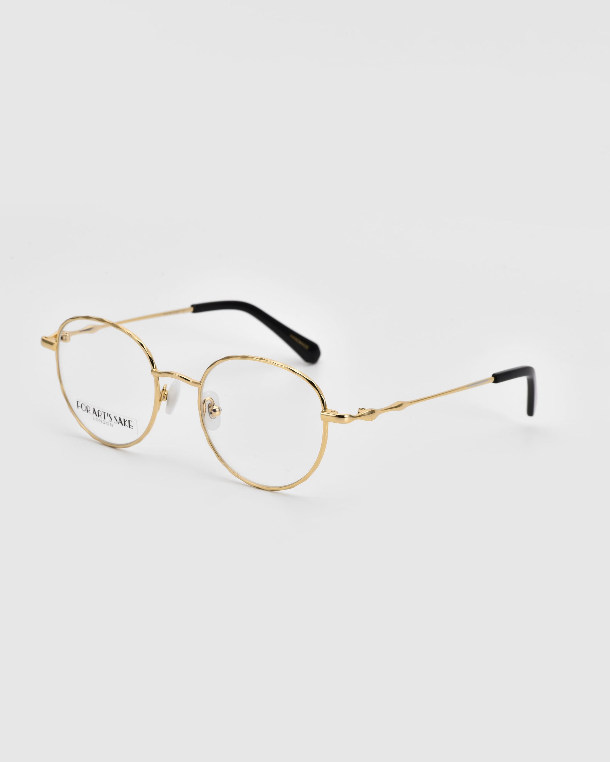 A pair of Bone I round, thin-framed eyeglasses from For Art&#39;s Sake® with luxurious metal detailing in gold-colored rims and black temple tips, positioned at an angle on a white background. The left lens is marked with &quot;FOR ART&#39;S SAKE&quot; in black text.