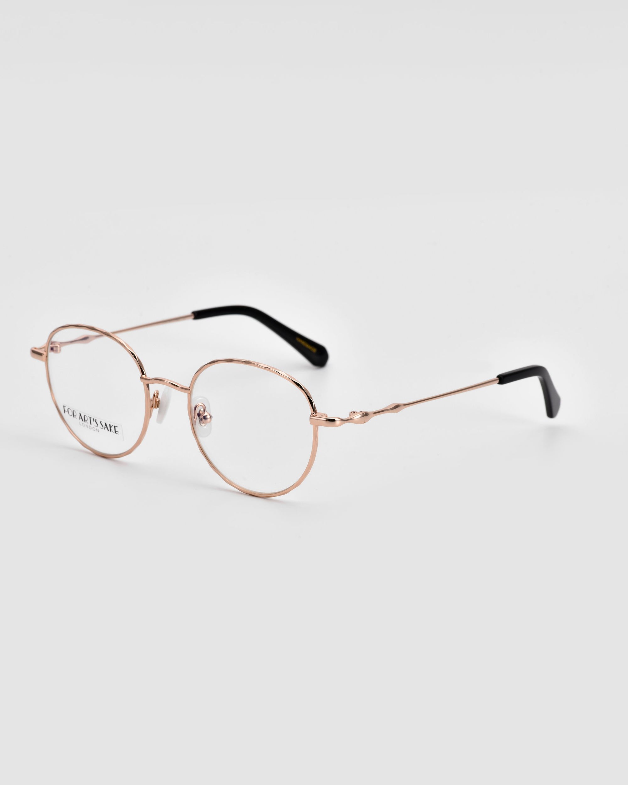 Delicate, gold-rimmed Bone I eyeglasses with oval lenses by For Art&#39;s Sake® feature &quot;JOC AETSNAE&quot; printed on one lens. The temples boast luxurious metal detailing and culminate in black tips. Set against a plain light grey background, these glasses exude sophistication.