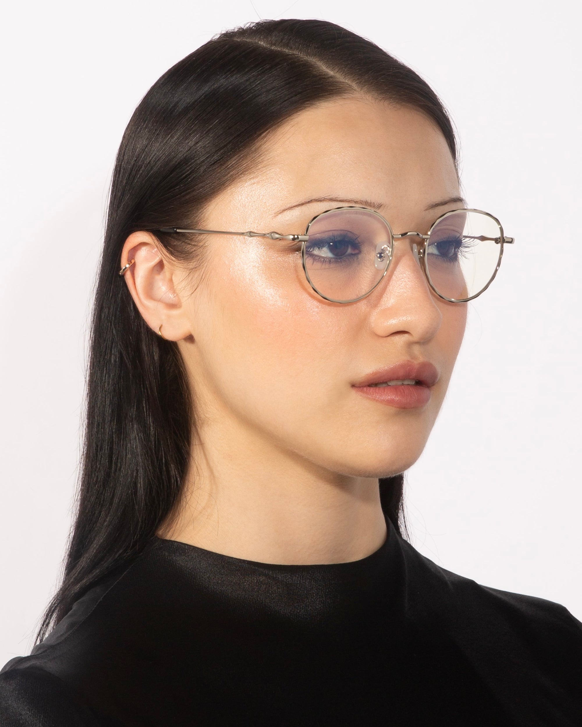 A person with long, straight dark hair and pale skin is wearing elegant eyewear from For Art&#39;s Sake®. Their Bone I round, thin-framed glasses feature real jade stone nose pads. They have a neutral expression and are shown against a plain white background.