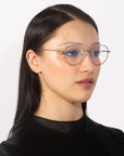 A person with long, straight dark hair and pale skin is wearing elegant eyewear from For Art's Sake®. Their Bone I round, thin-framed glasses feature real jade stone nose pads. They have a neutral expression and are shown against a plain white background.