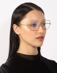 A woman with long, straight black hair wears elegant For Art's Sake® Bone II eyeglasses with clear lenses and jade nose pads. She has a subtle, natural makeup look and is dressed in a black top. The background is plain white.