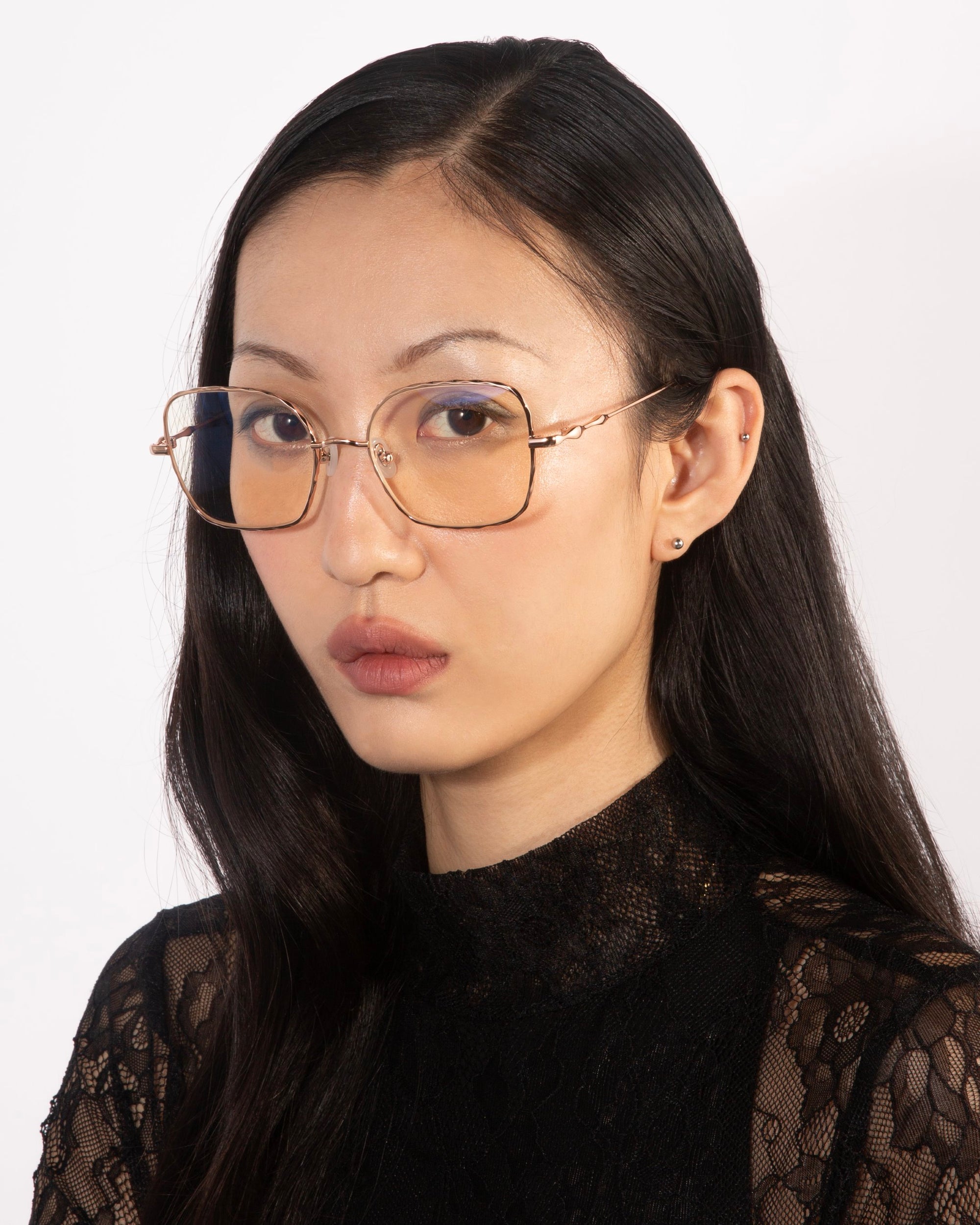 A woman with long black hair is wearing a black lace top and For Art&#39;s Sake® Bone I large square-framed glasses with a slight tint, featuring jade stone nose pads. She is looking slightly to the side with a serious expression. The background is light-colored and plain.