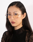 A woman with long black hair is wearing a black lace top and For Art's Sake® Bone I large square-framed glasses with a slight tint, featuring jade stone nose pads. She is looking slightly to the side with a serious expression. The background is light-colored and plain.