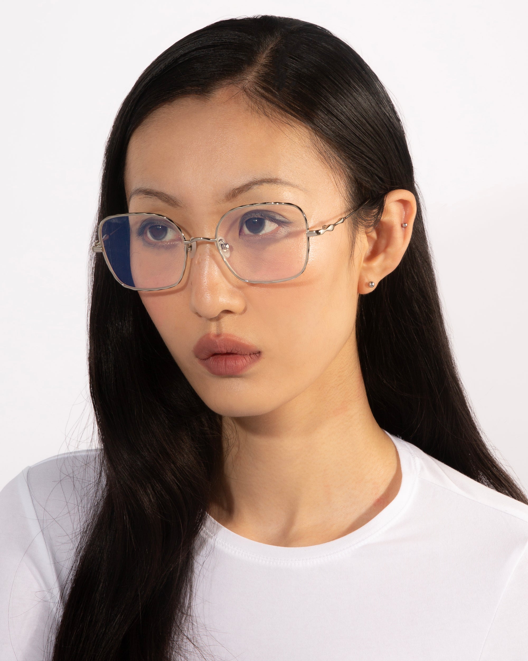 A person with long, straight black hair wears large, transparent-frame Bone II glasses from For Art's Sake® and a plain white top. The background is a simple white. The individual exudes refined elegance, with a neutral expression and looking slightly to the side.