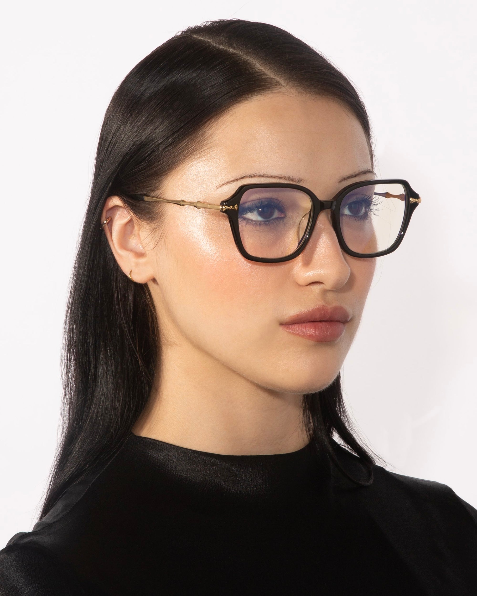 A woman with long, straight black hair is wearing the Delight eyeglasses by For Art's Sake®. The large, black-rimmed square frames feature gold detailing. She has a neutral expression and is shown against a white background while wearing a black top.