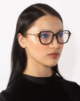 A woman with long, straight black hair is wearing the Delight eyeglasses by For Art's Sake®. The large, black-rimmed square frames feature gold detailing. She has a neutral expression and is shown against a white background while wearing a black top.