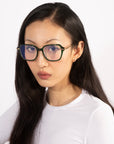 A person with long, dark hair wears Delight rectangular glasses with handcrafted frames from For Art's Sake®, paired with a white shirt, gazing directly at the camera against a plain white background.