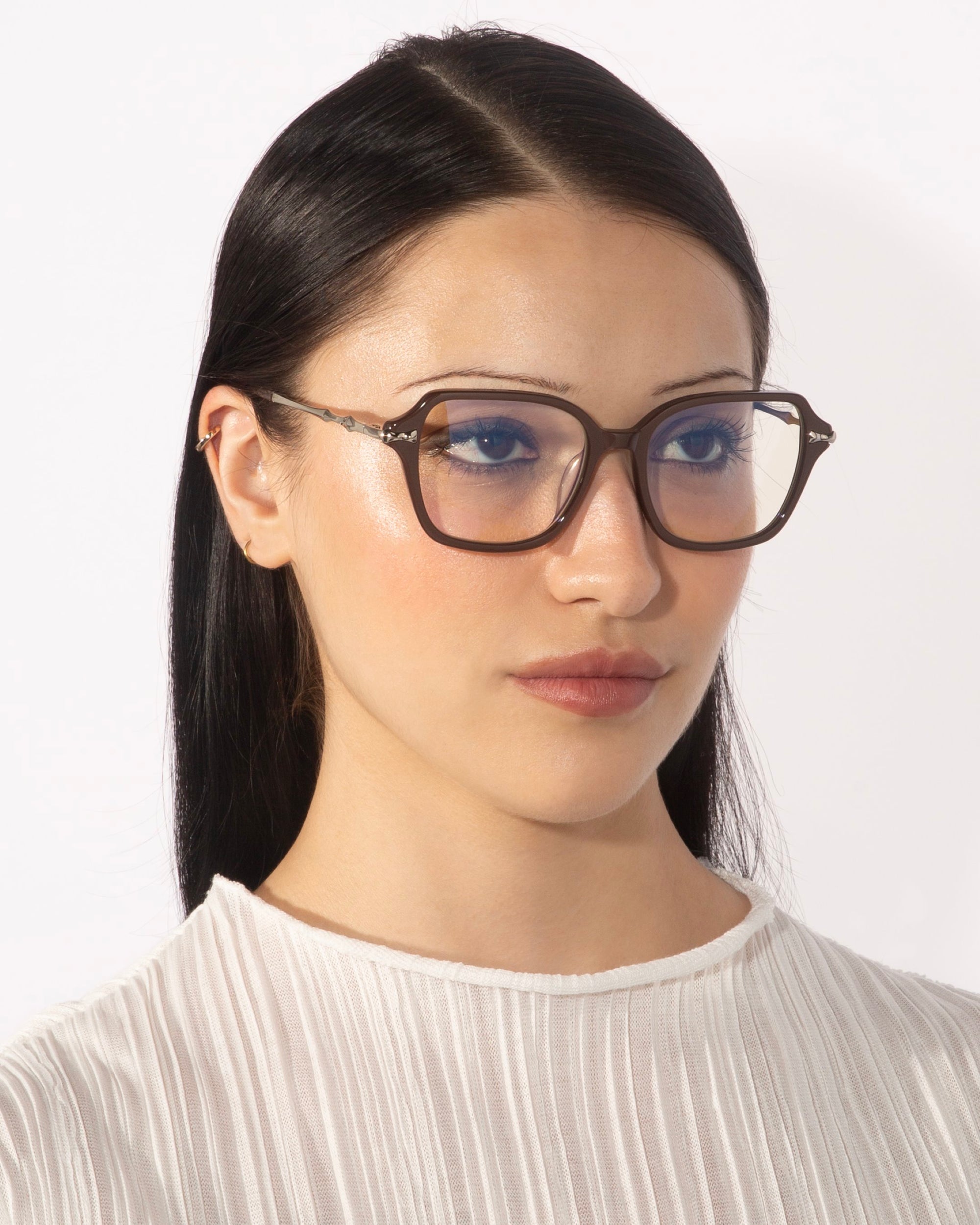 A person with long, straight dark hair wears large, angular square eyeglasses with brown frames from the Delight collection by For Art&#39;s Sake®. They have a neutral facial expression and are dressed in a white, textured top. The background is plain and white.