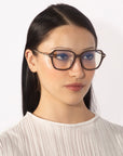 A person with long, straight dark hair wears large, angular square eyeglasses with brown frames from the Delight collection by For Art's Sake®. They have a neutral facial expression and are dressed in a white, textured top. The background is plain and white.