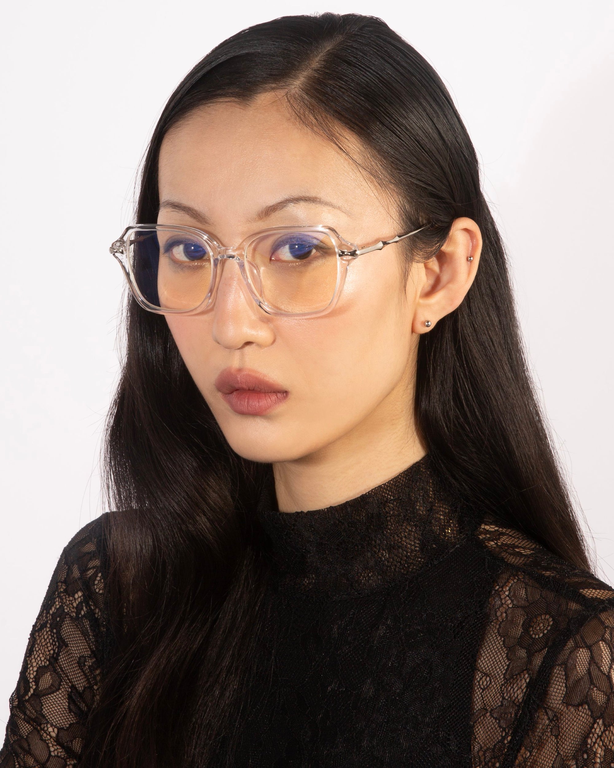 A woman with long, sleek black hair wears clear, oversized square eyeglasses with blue-tinted lenses from For Art's Sake®'s Delight collection. She has a calm, serious expression and is dressed in a black lace top. The plain, light background accentuates her vintage charm.