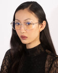 A woman with long, sleek black hair wears clear, oversized square eyeglasses with blue-tinted lenses from For Art's Sake®'s Delight collection. She has a calm, serious expression and is dressed in a black lace top. The plain, light background accentuates her vintage charm.