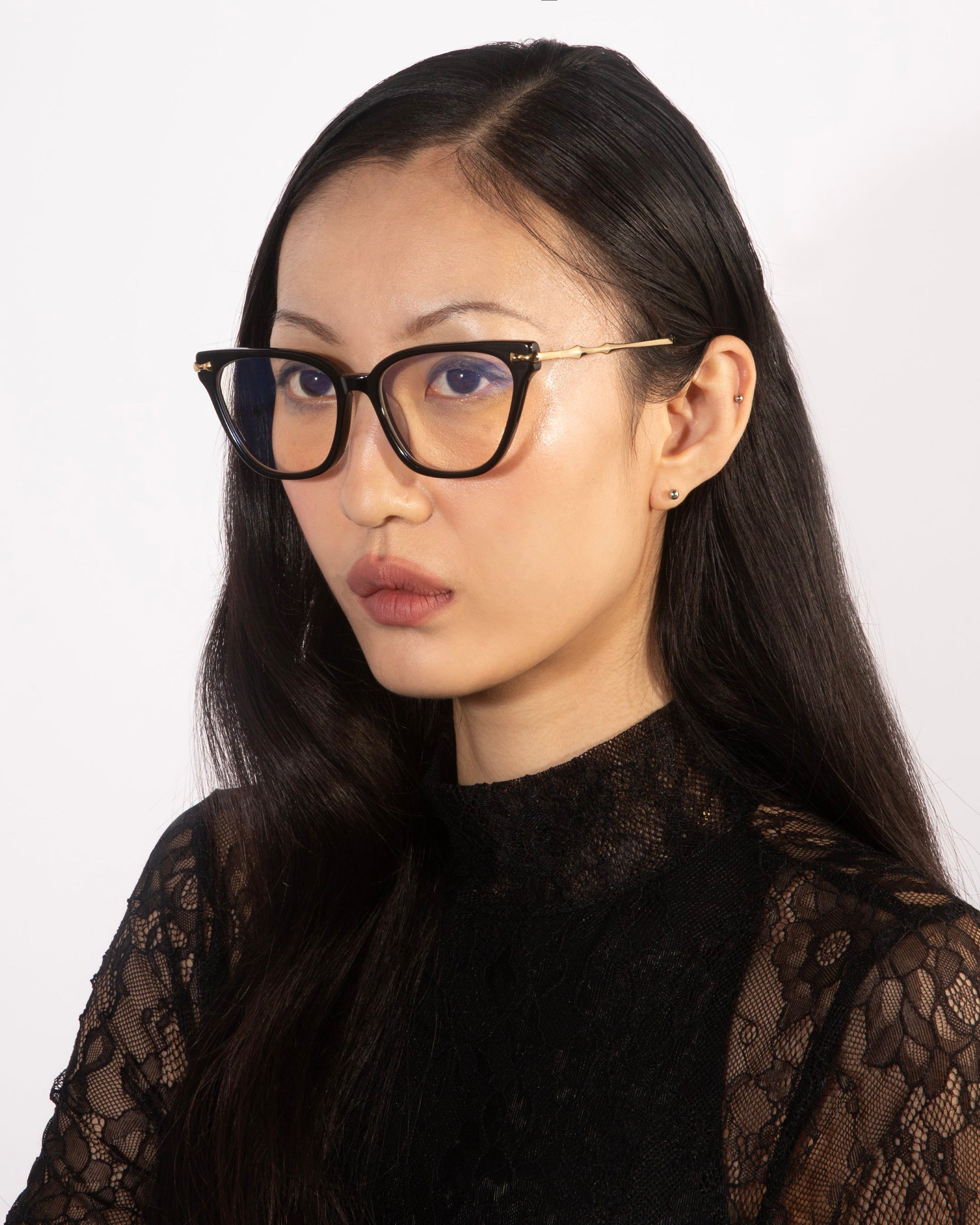 A woman with long, black hair wearing stylish Lexie cat-eye frames from For Art's Sake® and a black lace top poses for a portrait against a plain white background. She looks directly at the camera with a neutral expression, showcasing her eyeglasses.