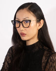 A woman with long, black hair wearing stylish Lexie cat-eye frames from For Art's Sake® and a black lace top poses for a portrait against a plain white background. She looks directly at the camera with a neutral expression, showcasing her eyeglasses.