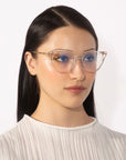 A person with long, straight dark hair is wearing the Lexie cat-eye frames by For Art's Sake® and a white pleated top. The background is plain white. The person has a neutral expression and is facing forward with a slight angle to the right.