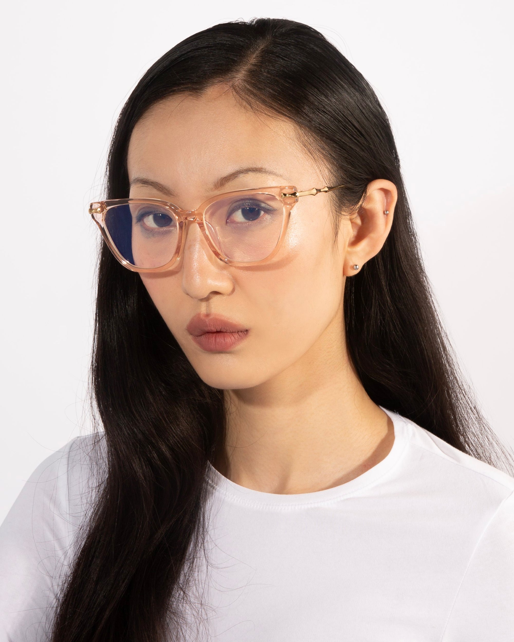 A person with long, dark hair is wearing large Lexie eyeglasses with transparent frames from For Art's Sake® and a white top. They are looking directly at the camera with a neutral expression against a plain white background.