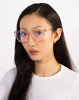 A person with long, dark hair is wearing large Lexie eyeglasses with transparent frames from For Art's Sake® and a white top. They are looking directly at the camera with a neutral expression against a plain white background.