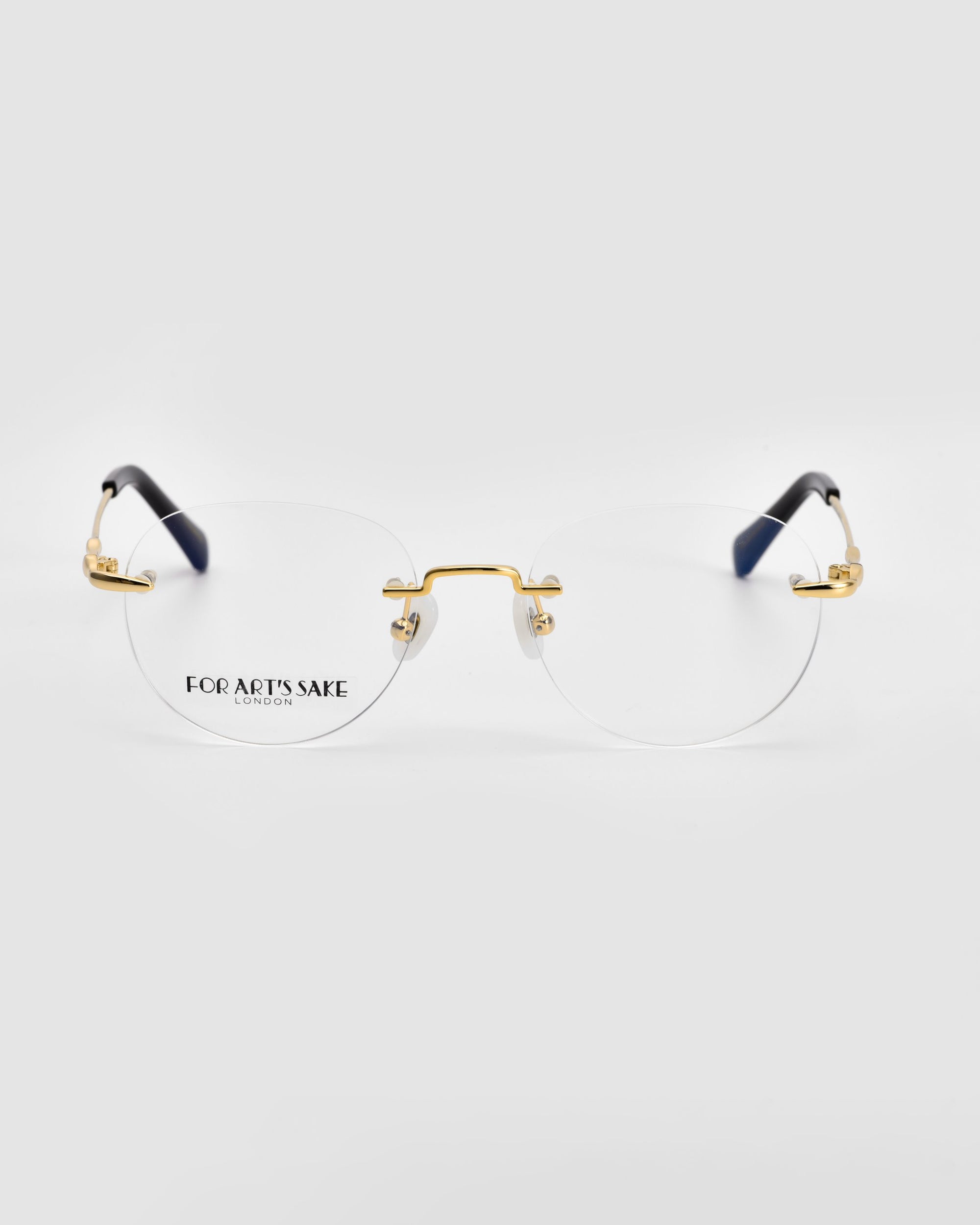 Introducing Misty, a pair of rimless eyeglasses featuring round lenses with a gold bridge and temples. The temple tips are coated in black. The left lens displays the brand name &quot;For Art&#39;s Sake®&quot; and &quot;London&quot; printed in black text. Set against a plain white background, these eyeglasses highlight their sleek wireframe construction.