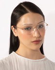 A young person with long, straight dark hair is wearing the Misty frameless round lenses by For Art's Sake® and a white textured blouse. The individual has a neutral expression and is looking slightly off-camera. The background is plain white.