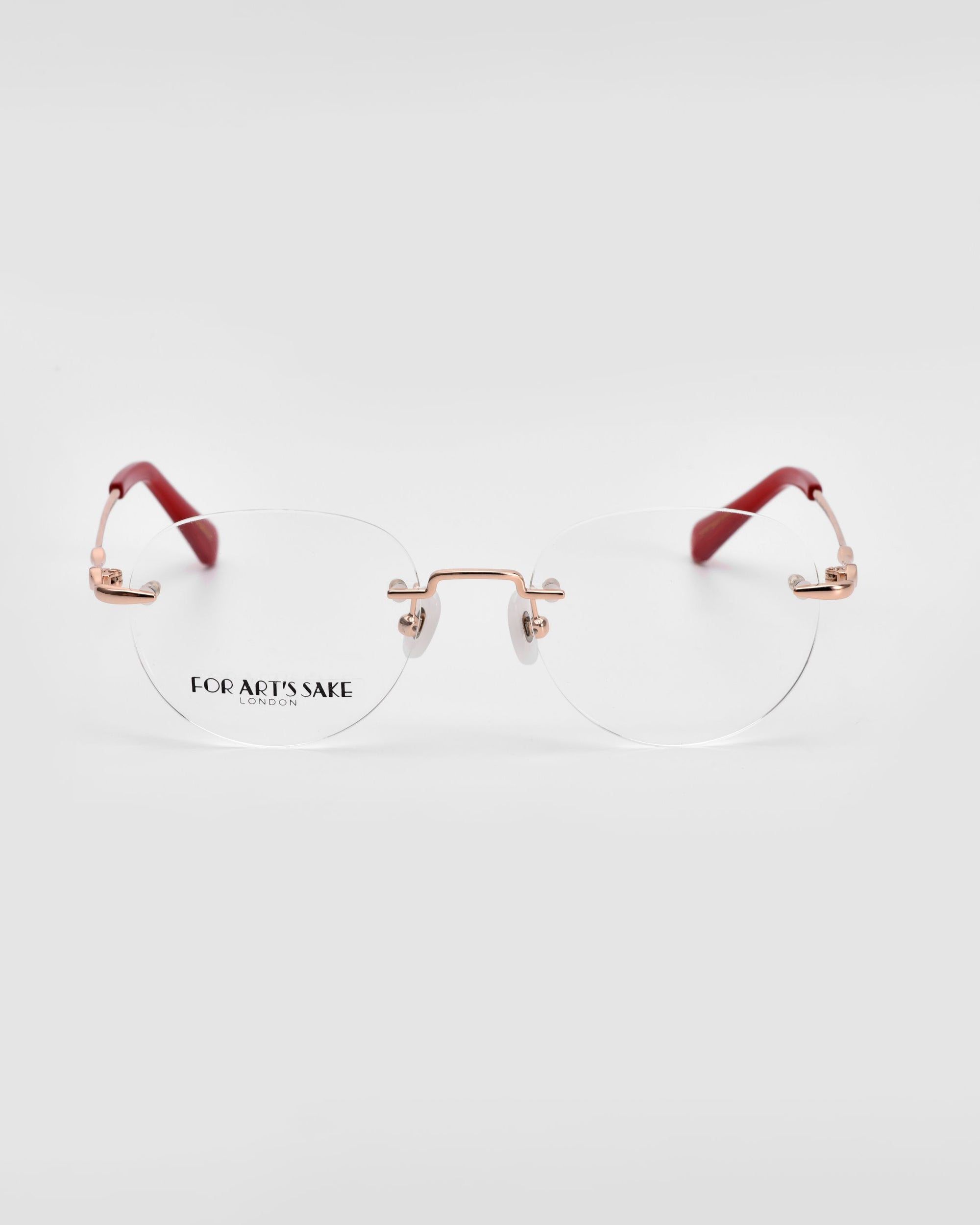 A pair of For Art&#39;s Sake® Misty rimless eyeglasses with rose gold metal accents and red temple tips. The clear round lenses have &quot;FOR ART&#39;S SAKE LONDON&quot; engraved on the lower left side. Featuring a sleek wireframe construction, the background is a plain light gray color.