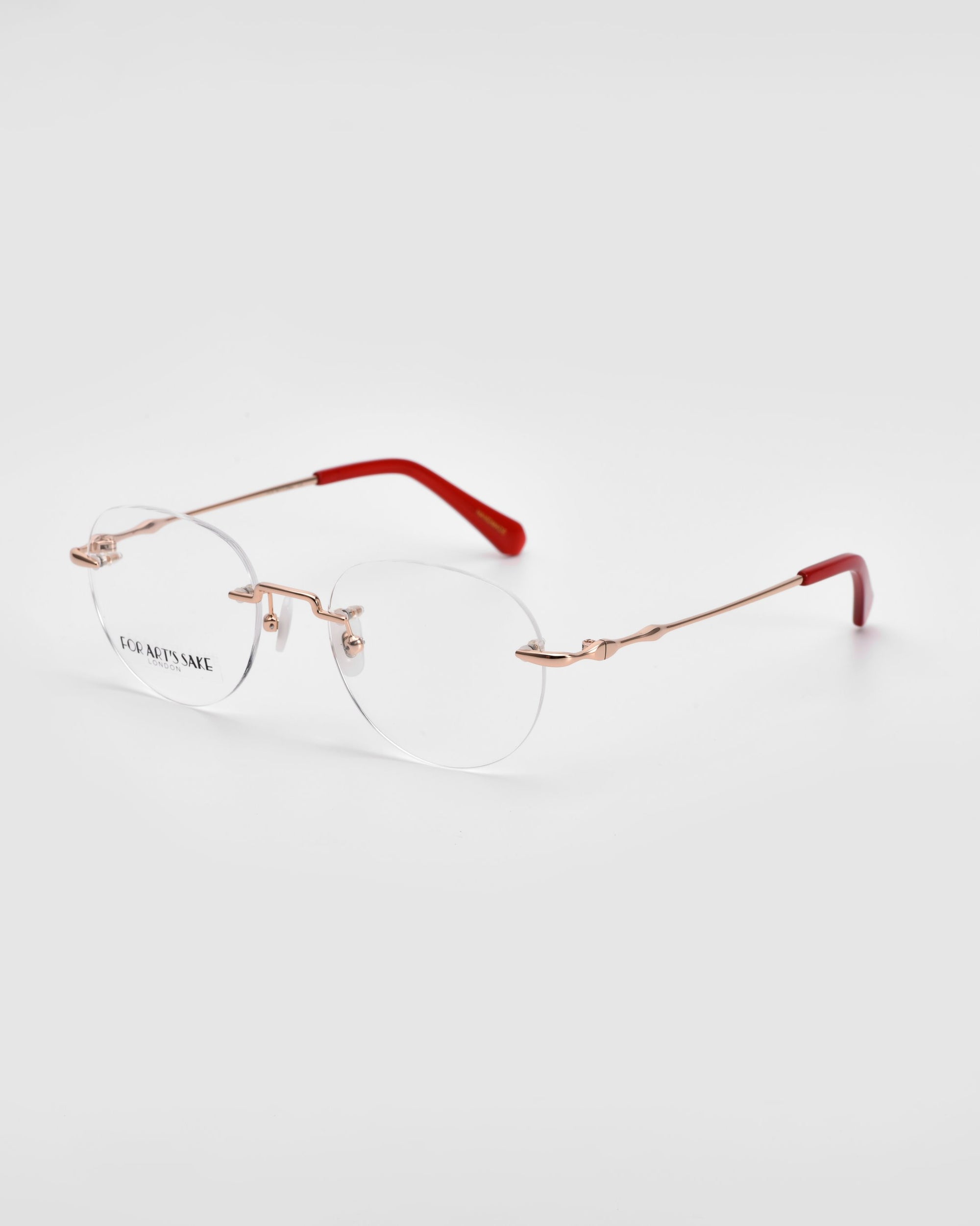 The Misty by For Art's Sake® features a pair of rimless eyeglasses with round lenses, gold-tone metal arms, and red tips on the ends of the temples. The minimalist and lightweight wireframe construction offers a clear and modern aesthetic against a white background.