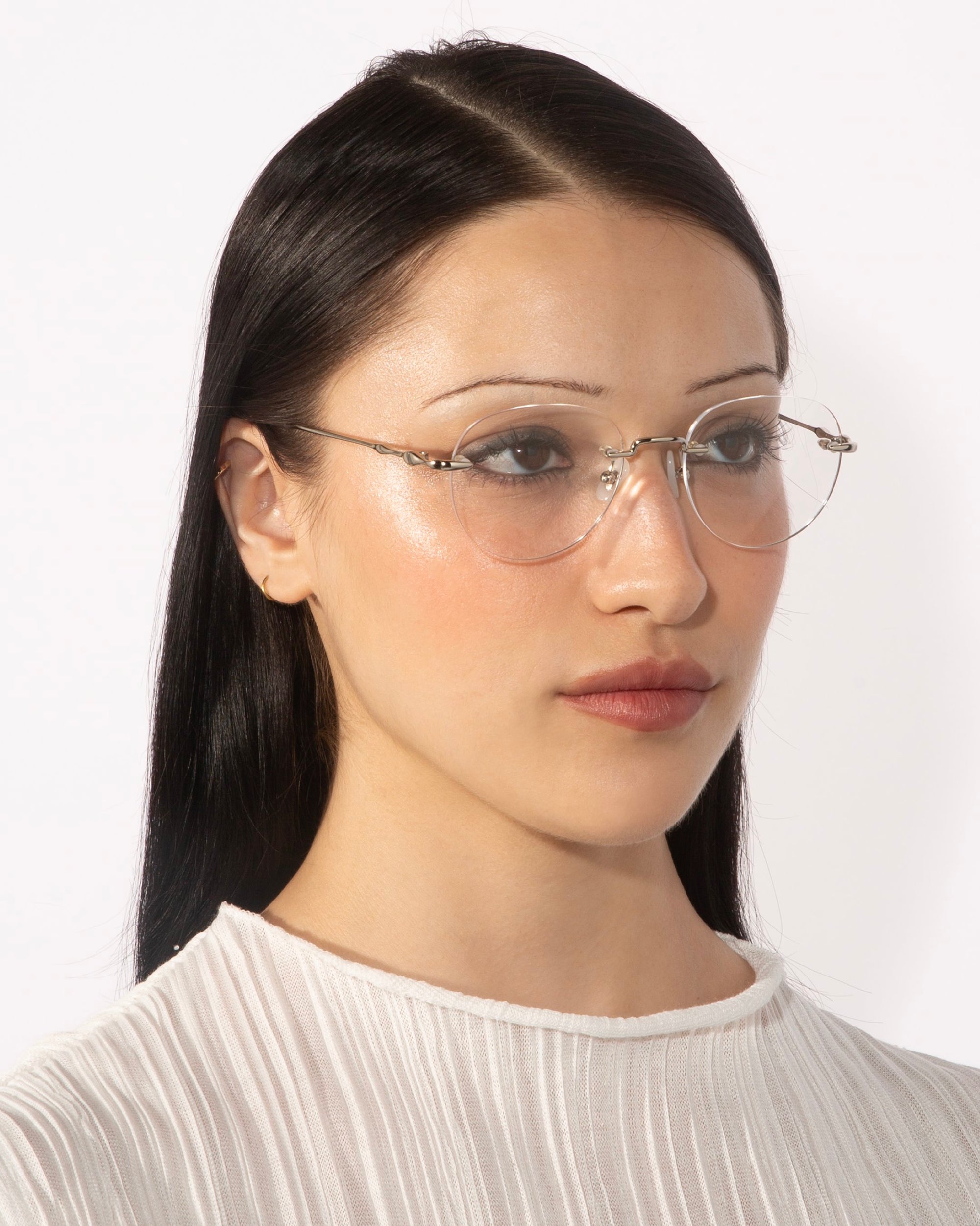 A person with straight black hair and clear, frameless round lenses, specifically the "Misty" model by For Art's Sake®, is shown from the shoulders up against a plain white background. They are wearing a white pleated top and have a neutral facial expression, looking slightly to the right.