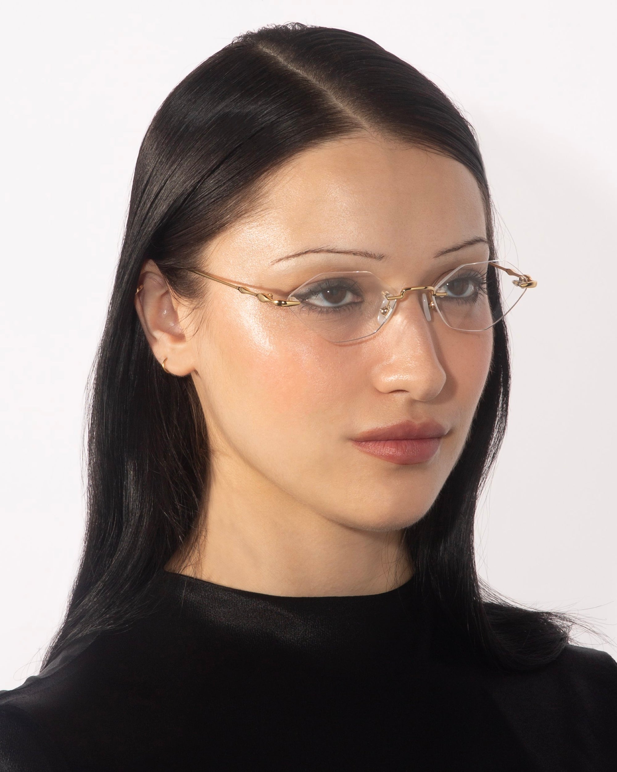 A person with long, straight black hair and light skin is wearing For Art's Sake® Zesty lightweight glasses with gold temples. They have a neutral expression and are dressed in a black top. The background is plain white.