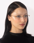 A person with long, straight black hair and light skin is wearing For Art's Sake® Zesty lightweight glasses with gold temples. They have a neutral expression and are dressed in a black top. The background is plain white.