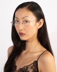 A woman with long black hair, wearing Zesty sleek thin square-shaped frameless structure glasses by For Art's Sake®, and a black lace camisole, faces the camera with a neutral expression. She has small earrings, and the background is plain white.