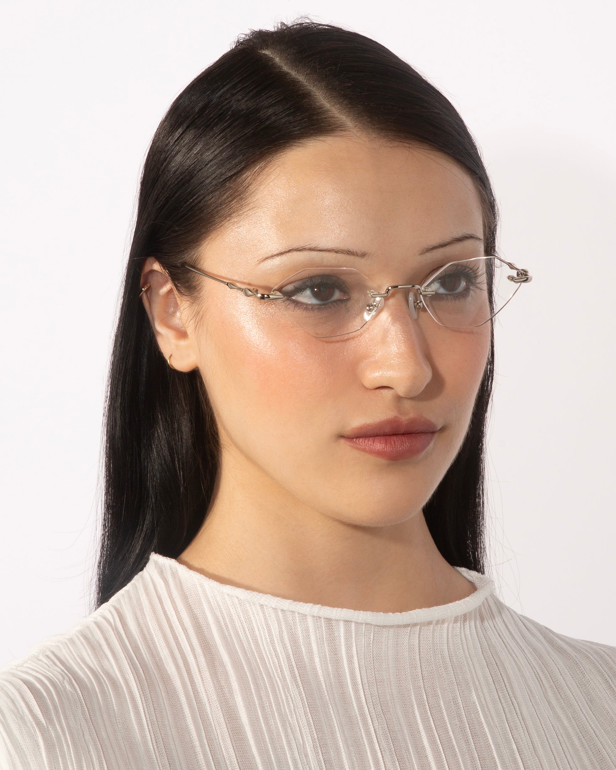 A woman with long dark hair, wearing Zesty lightweight, frameless glasses from For Art&#39;s Sake® and a white pleated blouse, looks slightly to the side against a plain white background.