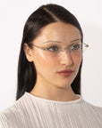 A woman with long dark hair, wearing Zesty lightweight, frameless glasses from For Art's Sake® and a white pleated blouse, looks slightly to the side against a plain white background.