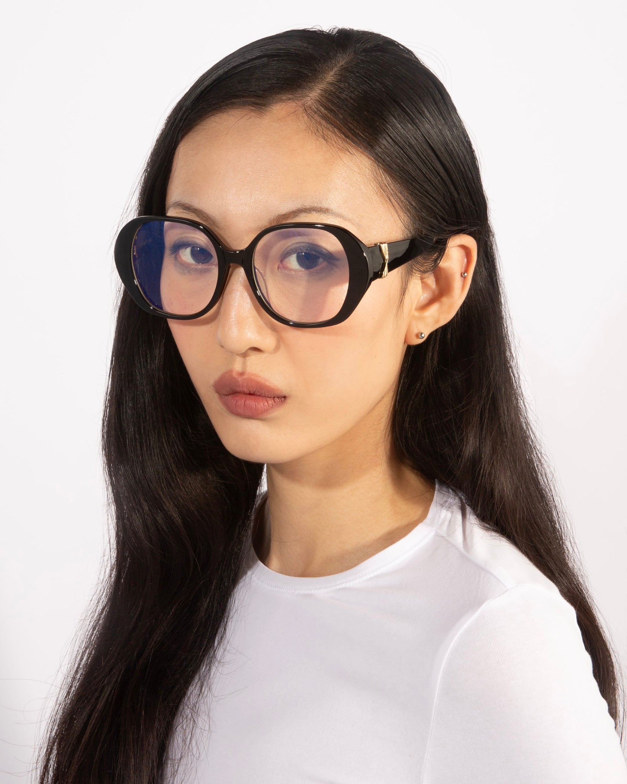 A person with long, dark hair dons the stylish and bold Bow glasses from For Art&#39;s Sake®, paired with a white top. The background is plain and light-colored, and their expression is neutral.