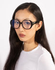 A person with long, dark hair dons the stylish and bold Bow glasses from For Art's Sake®, paired with a white top. The background is plain and light-colored, and their expression is neutral.