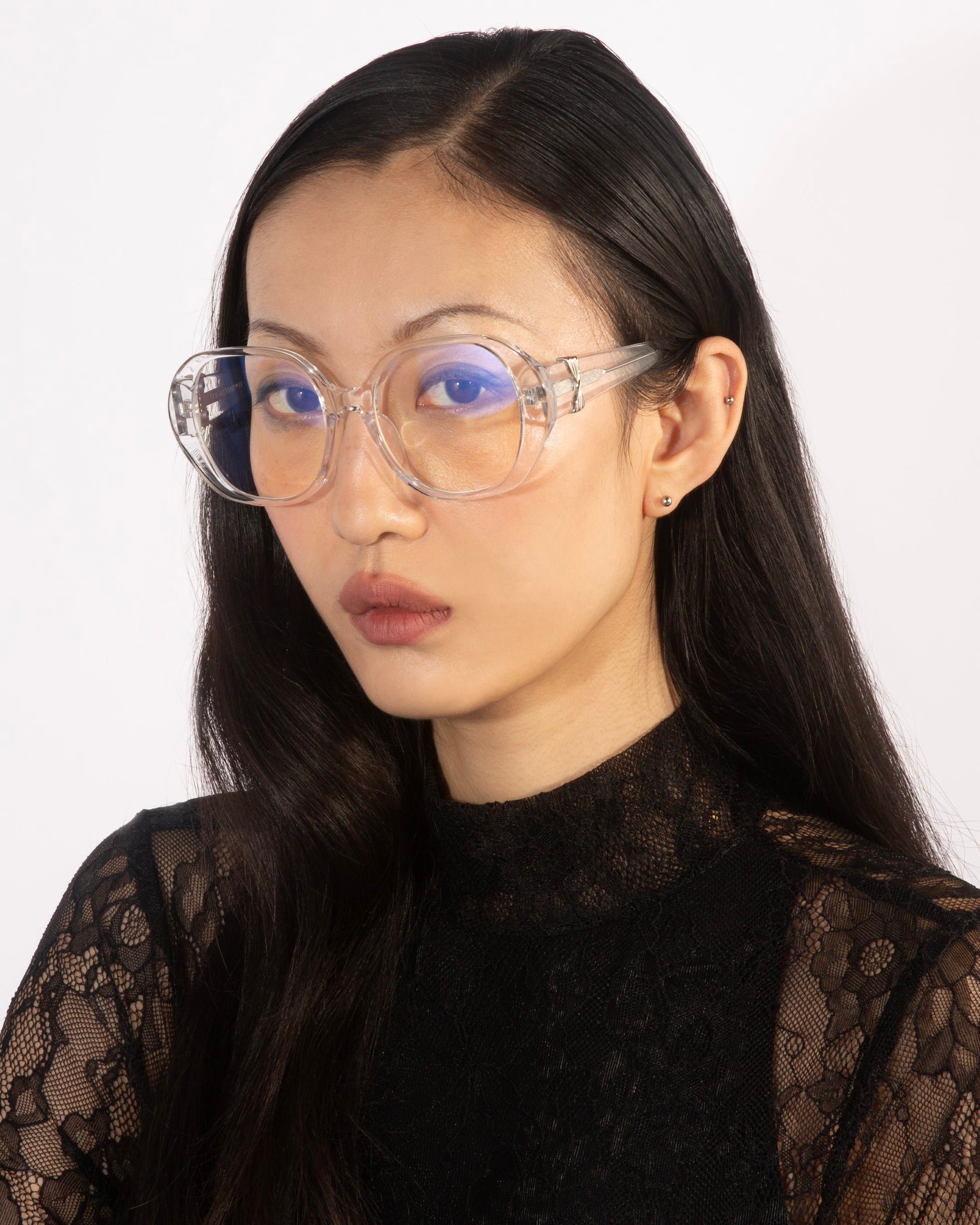 A woman with long black hair is wearing the Bow glasses from For Art's Sake®, which feature stylish, clear oversized lenses with a blue tint. She is dressed in a black lace top and gazes directly at the camera against a plain white background.