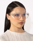 A person with long, straight black hair is wearing the For Art's Sake® Bow eyeglasses, which feature large, transparent lenses and a unique vintage design. The individual has a serious expression and is dressed in a white, textured high-neck top against a plain white background.