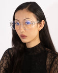 A woman with long black hair is wearing the Bow glasses from For Art's Sake®, which feature stylish, clear oversized lenses with a blue tint. She is dressed in a black lace top and gazes directly at the camera against a plain white background.