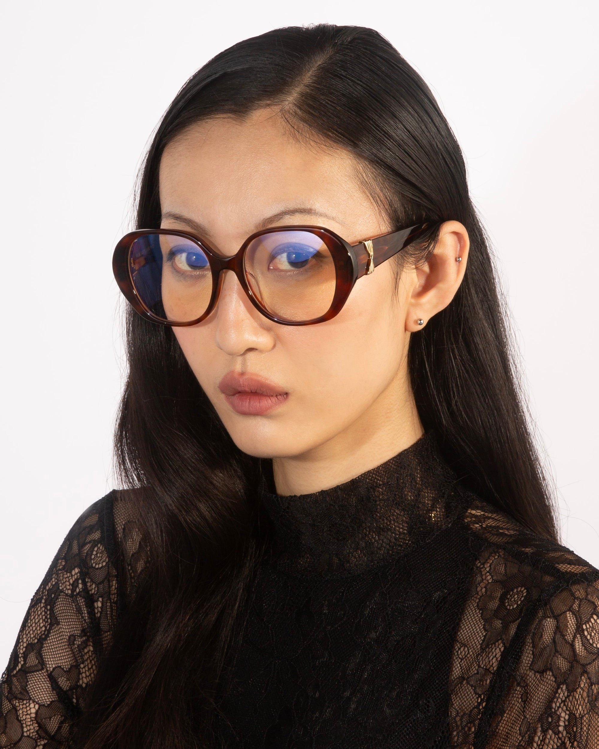 A woman with long, straight black hair is wearing the For Art's Sake® Bow optical glasses, featuring oversized lenses and thick, dark brown frames. She is dressed in a black lace top and posed against a plain white background. Her expression is neutral as she looks directly at the camera.