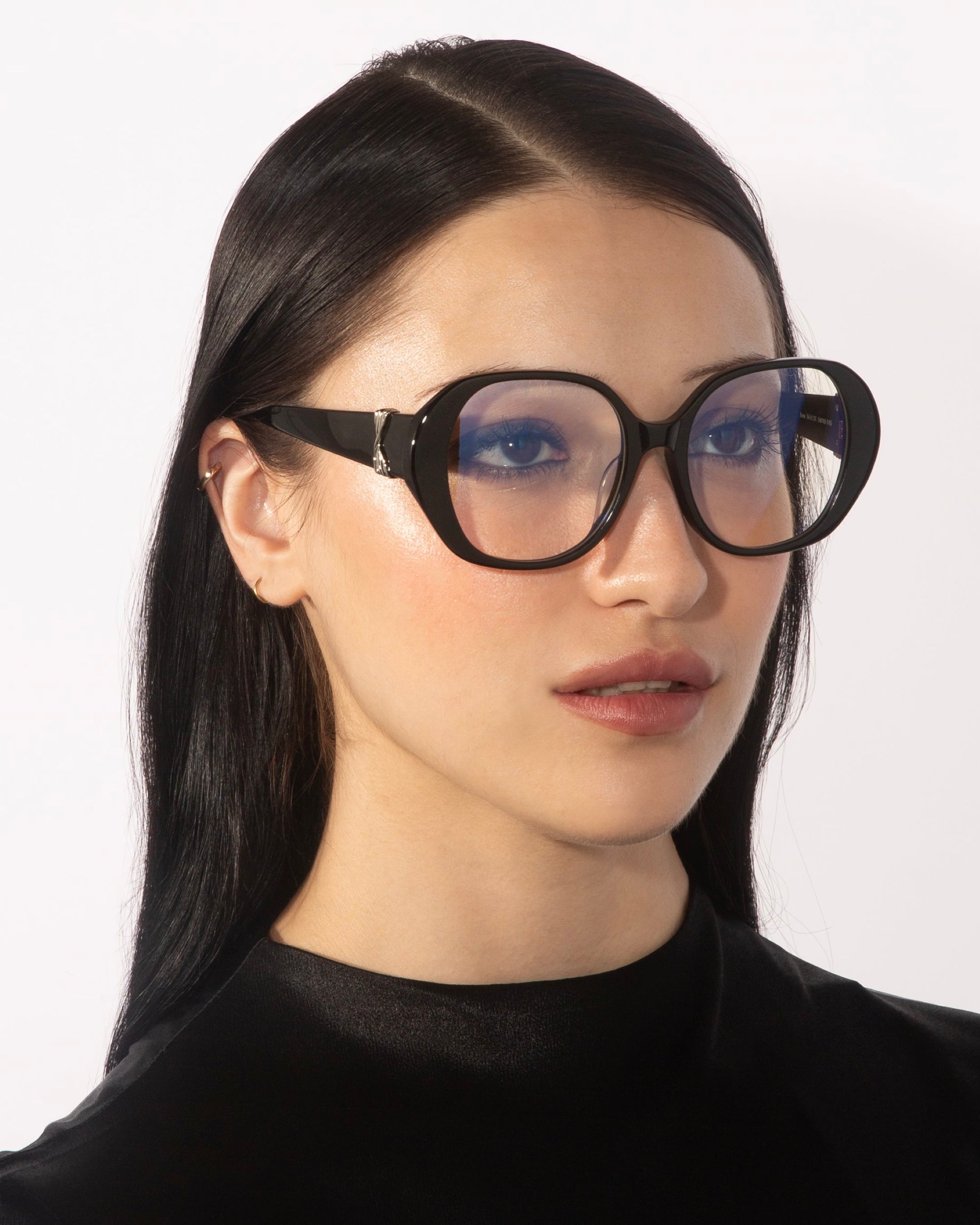 A person with long, straight black hair wears the Bow glasses by For Art's Sake®, which feature oversized lenses with a slight blue tint. They have a neutral expression and are dressed in a black top with a satin-like finish. The background is plain white.