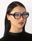 A person with long, straight black hair wears the Bow glasses by For Art's Sake®, which feature oversized lenses with a slight blue tint. They have a neutral expression and are dressed in a black top with a satin-like finish. The background is plain white.