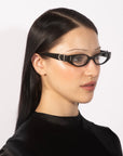 A person with long, straight black hair is wearing the Cigar narrow, black-framed glasses by For Art's Sake®, which give a contemporary edge. They are facing slightly to the left with a calm expression and dressed in a black high-neck top against a plain white background.